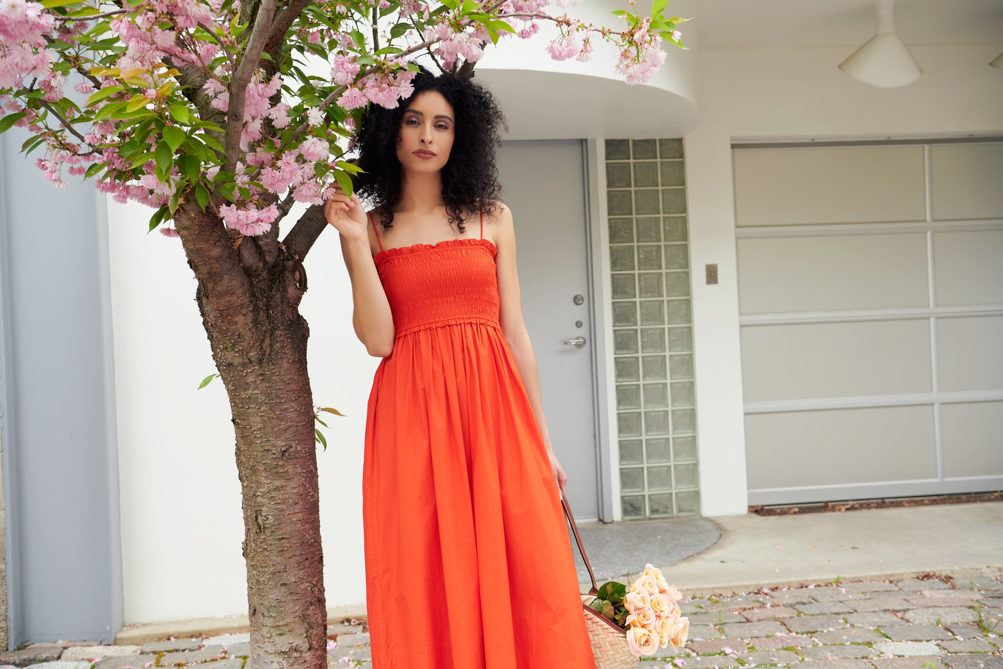 A woman stands beside a flowering tree wearing The Porta Smocked Spaghetti Dress by Apiece Apart