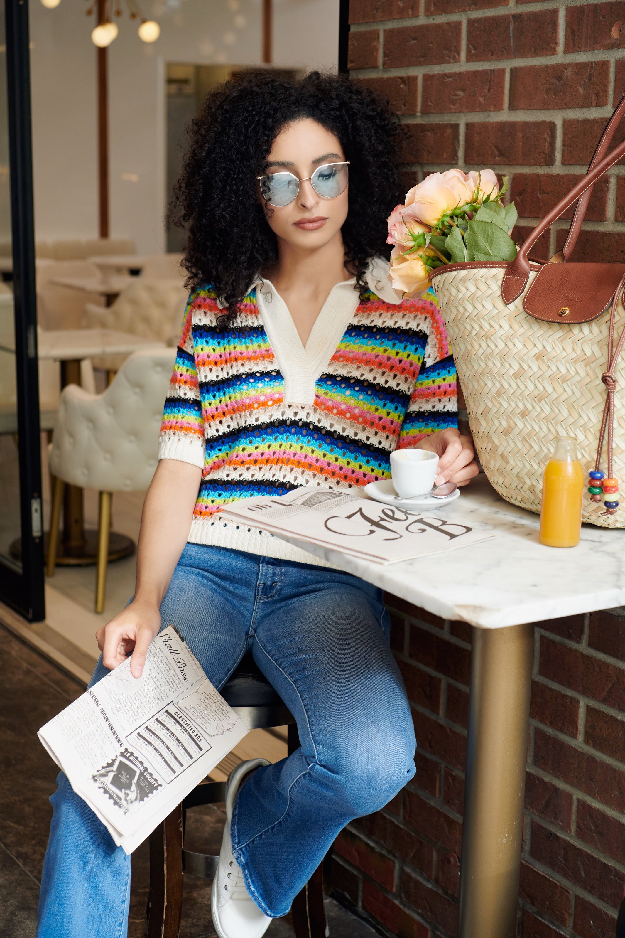 A woman sits outside a cafe with a newspaper, she is wearing the Nina Crochet-polo by Eleven Six and The Weekender jeans by MOTHER styled with Le Panier Pliage Tressé Handbag by Longchamp 