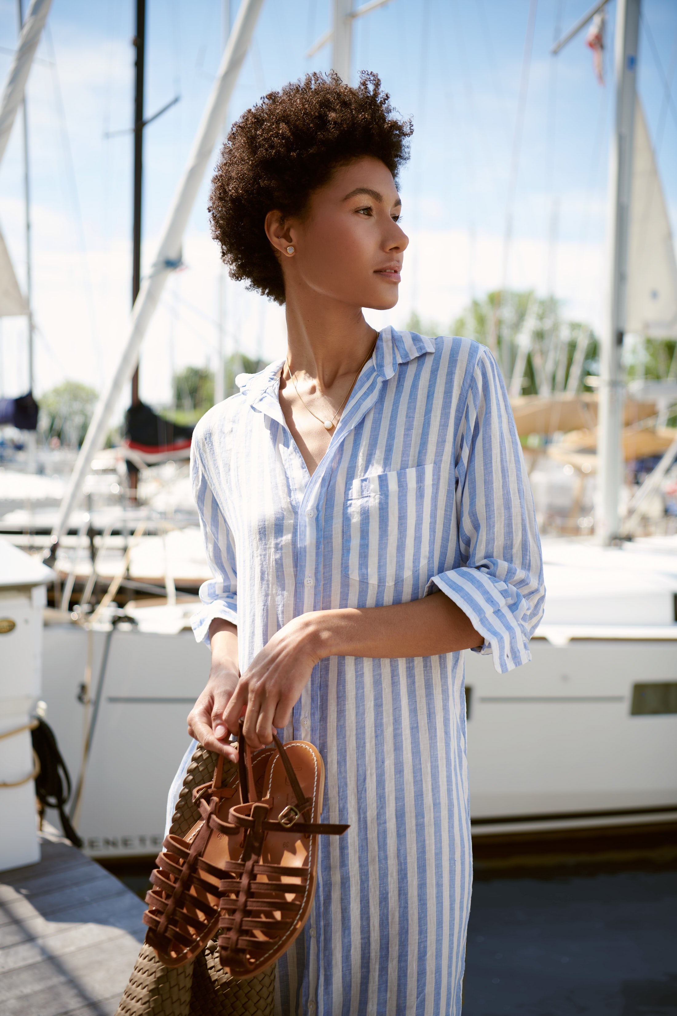 Woman stands on a dock beside a yacht wearing Frank & Eileen's Mary Shirt Dress while holding a Naghedi bag and K. Jacques sandals